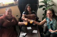 three women sitting around a table clinking their drinks together to cheer one another.