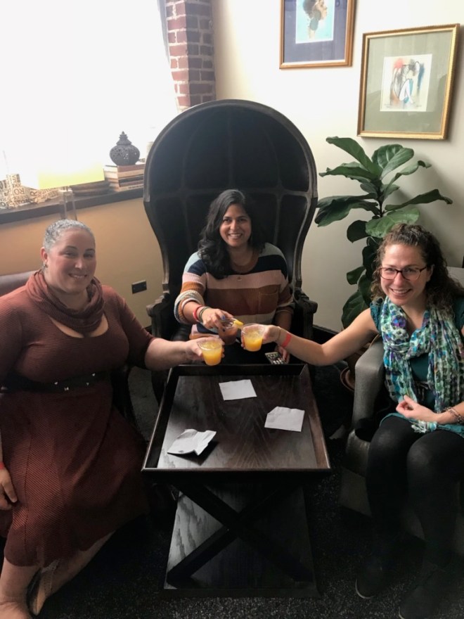three women sitting around a table clinking their drinks together to cheer one another.
